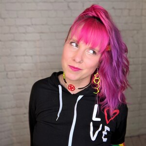 May include: A woman with pink and purple hair wearing a black hoodie with white drawstrings and the word "LOVE" written on it in white with a red heart. The hoodie has rainbow stripes on the sleeves.