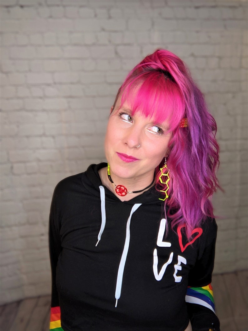 May include: A woman with pink and purple hair wearing a black hoodie with white drawstrings and the word "LOVE" written on it in white with a red heart. The hoodie has rainbow stripes on the sleeves.