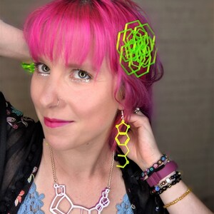May include: A woman with pink hair is wearing a white geometric necklace, neon green geometric earrings, and a neon green geometric hair clip. She is also wearing several bracelets on her wrist.