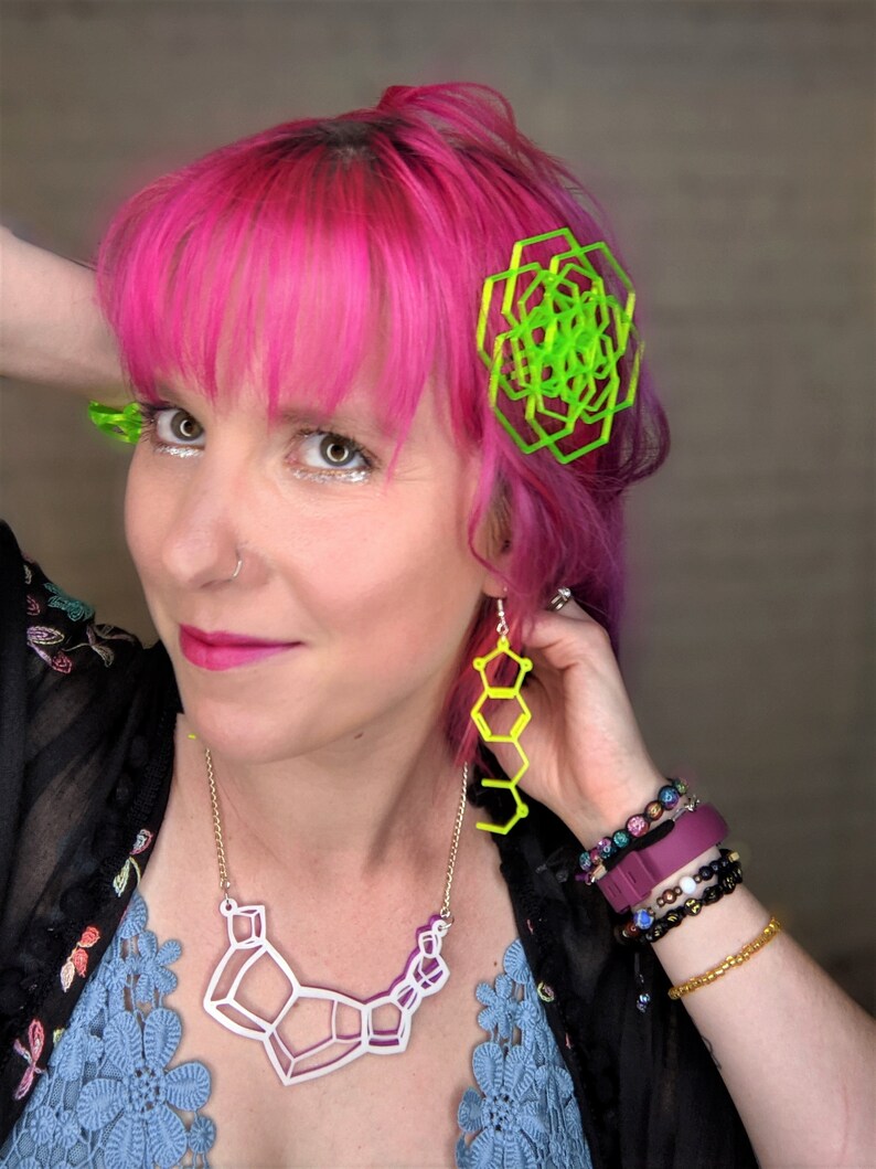 May include: A woman with pink hair is wearing a white geometric necklace, neon green geometric earrings, and a neon green geometric hair clip. She is also wearing several bracelets on her wrist.