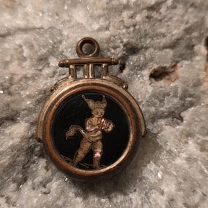 May include: Antique gold-toned pendant with a black face featuring a detailed illustration of a cherubic figure. The pendant has a circular design with a loop at the top for hanging. The cherub is depicted in gold and pink tones.