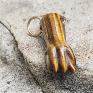 Puede incluir: Un colgante dorado con una piedra de ojo de tigre tallada en forma de mano con los dedos en forma de puño.
