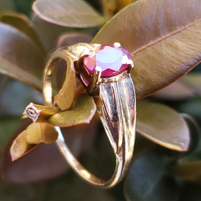 Puede incluir: Un anillo de oro con una gran gema de rub&iacute; de corte ovalado. La banda del anillo tiene un dise&ntilde;o detallado, y el rub&iacute; est&aacute; engastado en un engaste cl&aacute;sico de garras. El anillo se muestra sobre un fondo de hojas verdes y marrones.