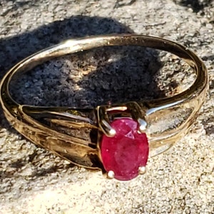 Puede incluir: Un anillo de oro con una gema ovalada de color rojo intenso. La banda del anillo tiene un dise&ntilde;o texturizado, y la gema est&aacute; sujeta por cuatro garras. El anillo se muestra sobre una superficie texturizada, a la luz del sol.