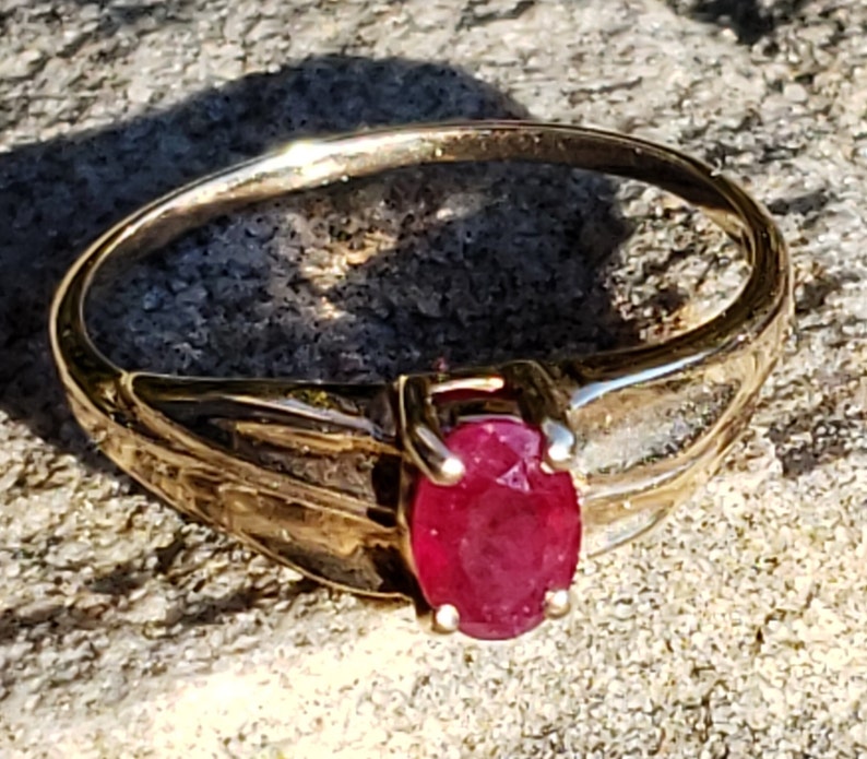 Puede incluir: Un anillo de oro con una gema ovalada de color rojo intenso. La banda del anillo tiene un dise&ntilde;o texturizado, y la gema est&aacute; sujeta por cuatro garras. El anillo se muestra sobre una superficie texturizada, a la luz del sol.