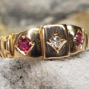 May include: A gold ring with three decorative sections. The outer sections feature a red gemstone in a diamond shape. The centre section has a diamond-shaped clear gemstone. The ring is on a textured surface.