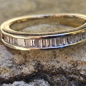 May include: A gold band ring with a row of rectangular-cut gemstones. The ring is set on a textured, light brown surface. The gemstones are clear and reflect light.