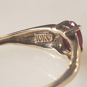 Puede incluir: Primer plano de un anillo de oro con una gema roja oscura grande, de forma ovalada. El anillo est&aacute; estampado con "10K" en el interior de la banda. El anillo est&aacute; sobre un fondo blanco.