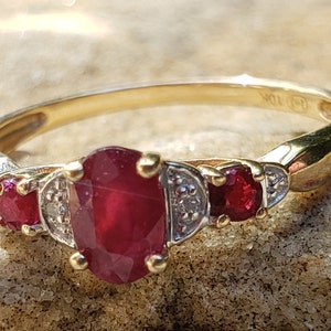 May include: A gold ring featuring three red rubies and two diamonds. The ring is set in a three-stone design with the largest ruby in the centre.