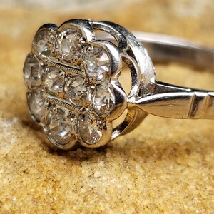 May include: A silver ring with a cluster of small, clear gemstones set in a floral design. The ring is on a brown, textured surface.