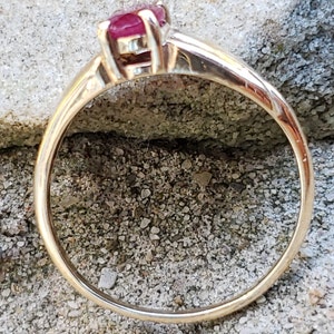 Puede incluir: Un anillo de oro con una vibrante gema roja. La banda del anillo es de oro pulido, y la gema est&aacute; engastada en un engaste cl&aacute;sico de cuatro puntas. El anillo se muestra sobre una superficie gris texturizada.