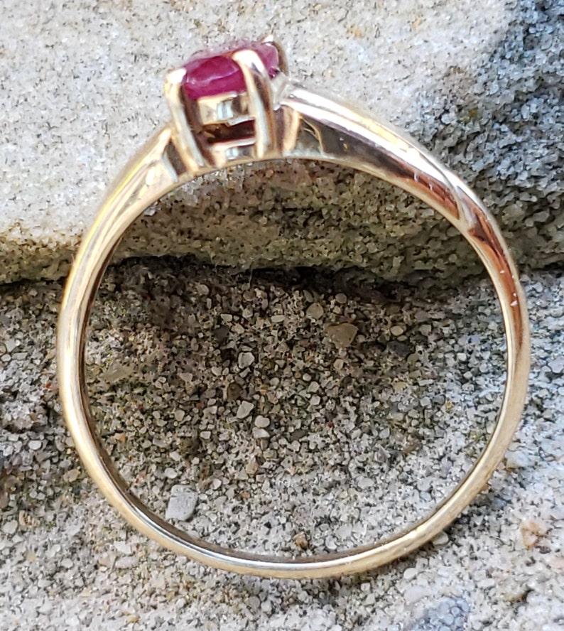Puede incluir: Un anillo de oro con una vibrante gema roja. La banda del anillo es de oro pulido, y la gema est&aacute; engastada en un engaste cl&aacute;sico de cuatro puntas. El anillo se muestra sobre una superficie gris texturizada.