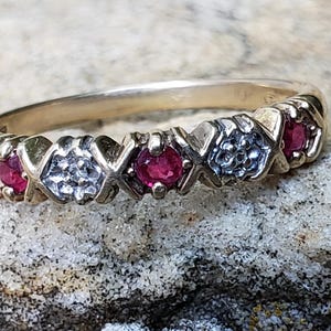 May include: A gold ring with alternating red gemstones and silver-coloured accents. The ring features a series of round, red stones set between decorative X and O shapes. The ring is set against a textured, light-coloured surface.