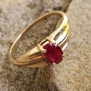 Puede incluir: Un anillo de oro con una gran gema de rub&iacute; de corte ovalado. La banda del anillo es lisa y pulida, con una ligera curva. El rub&iacute; es de color rojo intenso y el anillo est&aacute; sobre una superficie texturizada y clara.