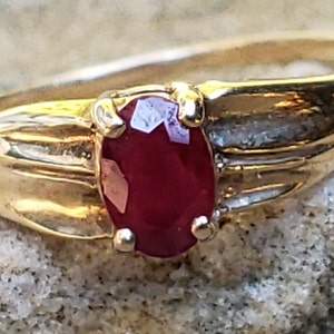 Puede incluir: Un anillo de oro con una gema de rub&iacute; rojo oscuro de corte ovalado. La banda del anillo tiene un dise&ntilde;o texturizado, y el rub&iacute; est&aacute; sujeto por cuatro garras. El anillo est&aacute; sobre un fondo neutro y texturizado.