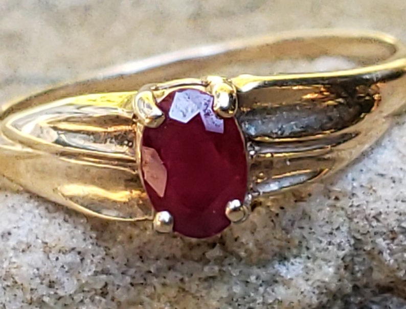 Puede incluir: Un anillo de oro con una gema de rub&iacute; rojo oscuro de corte ovalado. La banda del anillo tiene un dise&ntilde;o texturizado, y el rub&iacute; est&aacute; sujeto por cuatro garras. El anillo est&aacute; sobre un fondo neutro y texturizado.
