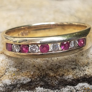 Könnte beinhalten: Ein goldener Bandring mit abwechselnd runden Rubinen und klaren Edelsteinen. Die Edelsteine sind in einer Kanalfassung gesetzt, wodurch eine durchgehende Farblinie um den Ring entsteht. Der Ring wird auf einer strukturierten Oberfläche präsentiert.