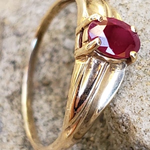 Puede incluir: Un anillo de oro con una gran gema de rub&iacute; de corte ovalado. El rub&iacute; es de color rojo intenso y est&aacute; engastado en un engaste de cuatro puntas. La banda del anillo tiene un dise&ntilde;o texturizado. El anillo se muestra sobre una superficie clara y texturizada.