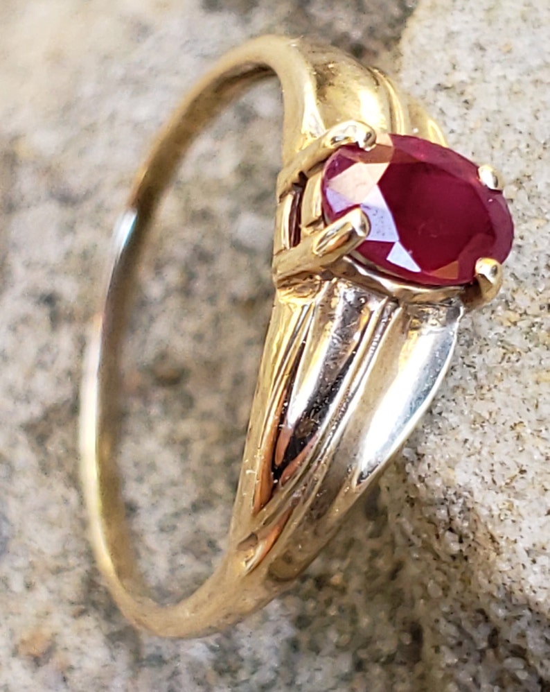 Puede incluir: Un anillo de oro con una gran gema de rub&iacute; de corte ovalado. El rub&iacute; es de color rojo intenso y est&aacute; engastado en un engaste de cuatro puntas. La banda del anillo tiene un dise&ntilde;o texturizado. El anillo se muestra sobre una superficie clara y texturizada.