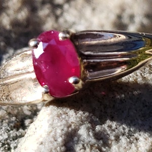 Puede incluir: Un anillo de oro con una gran gema de rub&iacute; de corte ovalado. El rub&iacute; es de color rojo vibrante y est&aacute; engastado en una banda de oro con un dise&ntilde;o detallado en los laterales. El anillo se muestra sobre una superficie texturizada de color claro.