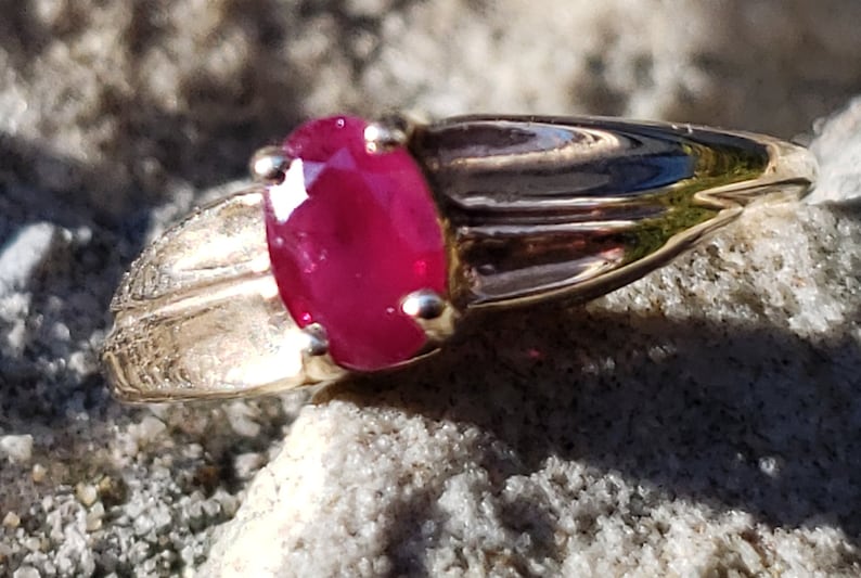 Puede incluir: Un anillo de oro con una gran gema de rub&iacute; de corte ovalado. El rub&iacute; es de color rojo vibrante y est&aacute; engastado en una banda de oro con un dise&ntilde;o detallado en los laterales. El anillo se muestra sobre una superficie texturizada de color claro.