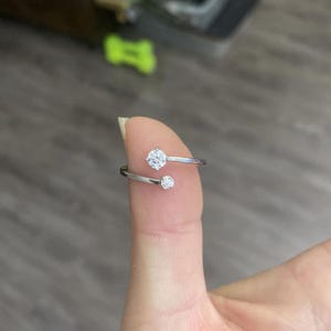 Op de afbeelding: Een zilverkleurige ring met twee heldere, ronde edelstenen. De ring heeft een open ontwerp, met een edelsteen aan de bovenkant en een kleinere eronder. De ring wordt op een vinger getoond.