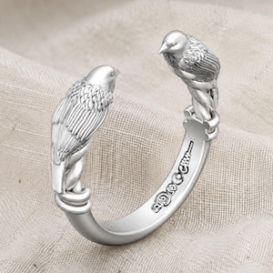 May include: A silver-toned open cuff ring featuring two detailed bird figures facing each other at the ends. The band is engraved with a series of small, circular decorative symbols along the outer side, showcasing a polished metallic finish.