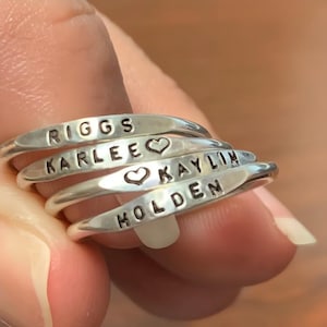 Peut inclure: Plusieurs bagues en argent empilées, chacune gravée d'un nom. Les noms incluent "RIGGS", "KARLEE" avec un symbole de cœur, "KAYLIN" avec un symbole de cœur et "HOLDEN". Les bagues sont empilées sur un doigt.