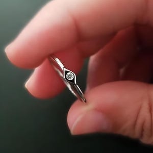 以下が含まれることがあります： 小さなダイヤモンドのような石が、目の形をしたベゼルにセットされたシルバーリング。リングは2本の指の間に挟まれ、繊細なデザインとミニマリストスタイルを際立たせています。バンドは細く、磨かれています。