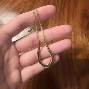 May include: A gold-coloured necklace featuring a teardrop pendant. The pendant has a smooth, curved form, suspended from a delicate chain. The necklace is held in a hand against a wooden background.