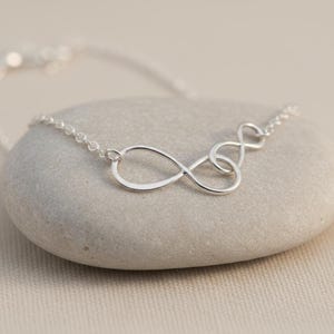 May include: A silver necklace with three intertwined infinity symbols. The necklace is shown on a grey stone, with the chain extending out of view. The design is simple and elegant, suitable for everyday wear.