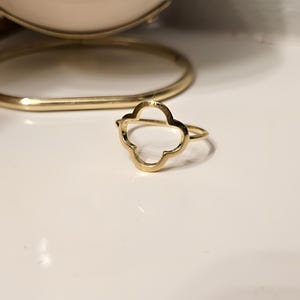 Puede incluir: Un anillo dorado con un diseño de trébol de cuatro hojas. El anillo es de metal brillante y descansa sobre una superficie blanca. Una joya delicada.