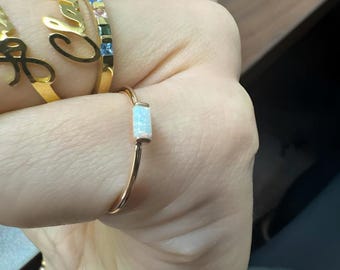 Puede incluir: Primer plano de una mano con varios anillos de oro. Destaca un anillo de oro rosa con una piedra de ópalo rectangular. Otros anillos presentan letras escritas y piedras preciosas de colores.