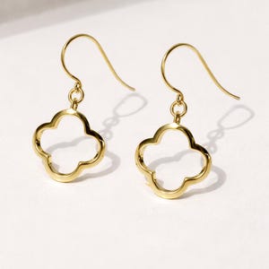 Peut inclure: Une paire de boucles d'oreilles pendantes dorées en forme de trèfle à quatre feuilles. Les boucles d'oreilles ont une fermeture à crochet et sont présentées sur une surface blanche.