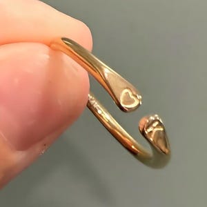 Puede incluir: Un anillo dorado con un diseño en forma de corazón. El anillo tiene un diseño abierto con un recorte en forma de corazón en un extremo. El anillo está sostenido por los dedos de una persona.