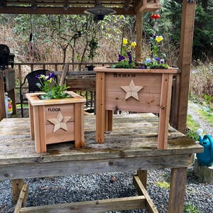 May include: Two wooden planters with star cutouts, one labeled "FLOOR" and the other "12 X 18 X 18". Both are filled with colourful flowers. The planters are on a weathered wooden table, suggesting outdoor use.