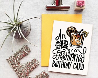 以下が含まれることがあります： 「An Old Fashioned Birthday Card」という黒い文字が書かれた白いカード。文字の上に、チェリーとレモンのスライスが添えられた、オールドファッションの漫画イラストが描かれている。