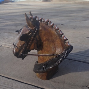 Puede incluir: Una escultura de bronce de la cabeza de un caballo con una brida de cuero marr&oacute;n. La escultura est&aacute; sobre una superficie de madera.