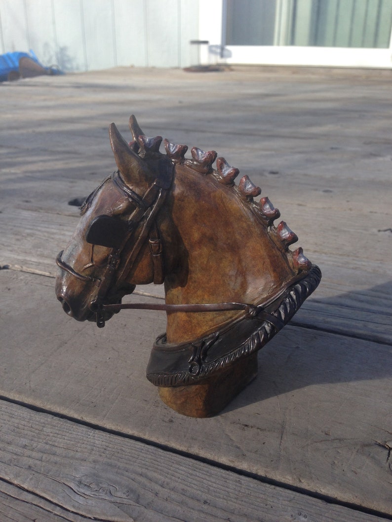 Puede incluir: Una escultura de bronce de la cabeza de un caballo con una brida de cuero marr&oacute;n. La escultura est&aacute; sobre una superficie de madera.