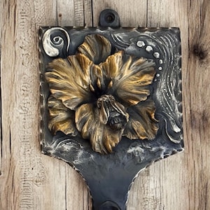 May include: A decorative metal wall hook with a floral design. The hook features a bronze-colored flower with detailed petals set against a dark gray background. The hook is approximately 6 inches tall.