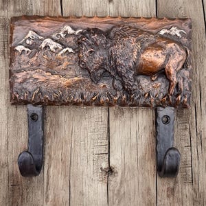 May include: A rustic wall-mounted coat rack featuring a detailed relief of a bison and mountain range. The bison is rendered in shades of brown and copper, with black metal hooks below. The rack is mounted on a wooden background.
