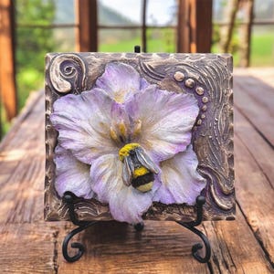 May include: A square, textured, decorative tile featuring a large, detailed flower in shades of purple and white, with a yellow-and-black bumblebee. The tile is set on a black metal stand, showcasing the floral relief against a brown background.