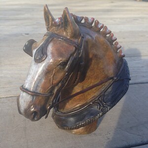 Puede incluir: Una escultura de bronce de la cabeza de un caballo con una brida de cuero marr&oacute;n. El caballo tiene una mancha blanca en la cara y una crin roja.