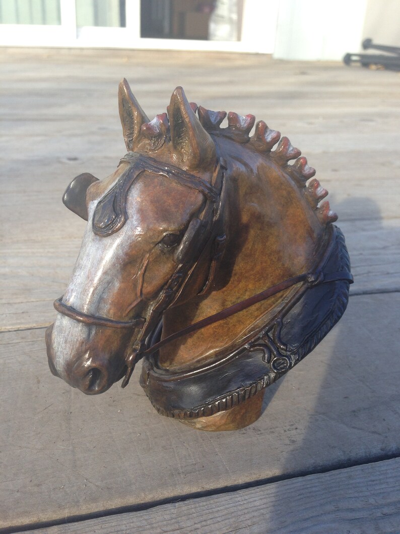 Puede incluir: Una escultura de bronce de la cabeza de un caballo con una brida de cuero marr&oacute;n. El caballo tiene una mancha blanca en la cara y una crin roja.