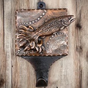 May include: A bronze-colored metal stocking holder featuring a detailed relief of a fish leaping upwards. The fish is set against a rectangular plaque with decorative wave and dot patterns. A hook is attached below for hanging. The background is a wooden surface.