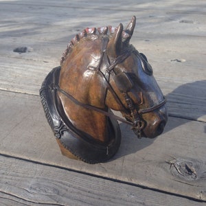 Puede incluir: Una escultura de bronce de la cabeza de un caballo con una brida de cuero. La crin del caballo est&aacute; esculpida con detalle y la escultura est&aacute; montada sobre una base de madera.