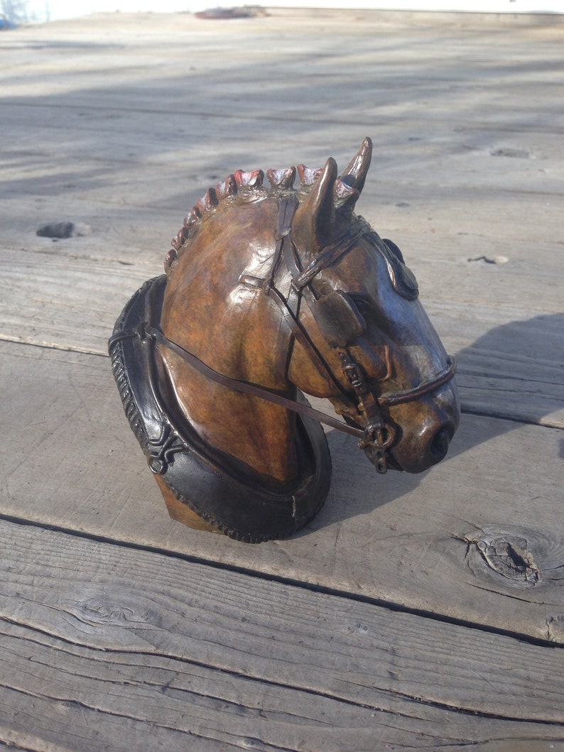 Puede incluir: Una escultura de bronce de la cabeza de un caballo con una brida de cuero. La crin del caballo est&aacute; esculpida con detalle y la escultura est&aacute; montada sobre una base de madera.