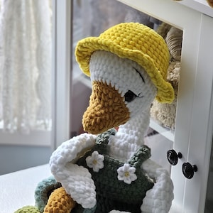 May include: A crocheted goose toy wearing a yellow hat and green overalls with white flower accents. The goose is sitting on a white fluffy rug.