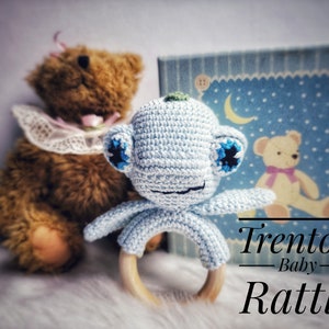 May include: A light blue crocheted baby rattle with a wooden ring. The rattle is shaped like a small animal with blue eyes and a smiling face. The text "Trenton Baby Rattle" is visible in the image.