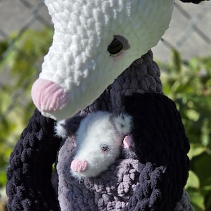 May include: A handmade crochet opossum plush toy with a baby opossum nestled in its arms. The large opossum is primarily gray, white, and black, with a pink nose and paws. The baby opossum is white with gray markings and a pink nose.